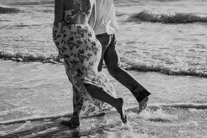 A black and white image shows two people walking in the surf at the beach with waves splashing at their feet.