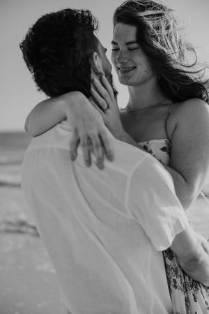 A romantic black and white portrait of a couple embracing on the beach with windswept hair and joyful expressions.