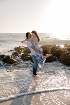 A romantic couple embraces on rocky beach at sunset while waves crash around their feet.