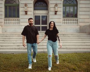 A couple in casual black shirts and blue jeans holds hands while standing on stone steps of a historic building.