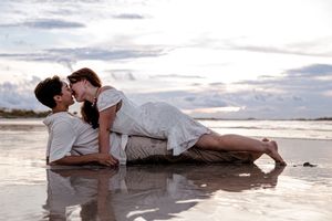 Romantic couple in white attire lying together on wet sand at beach during cloudy sunset.
