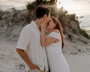 Romantic couple in white clothing share intimate moments on a sandy beach during sunset.