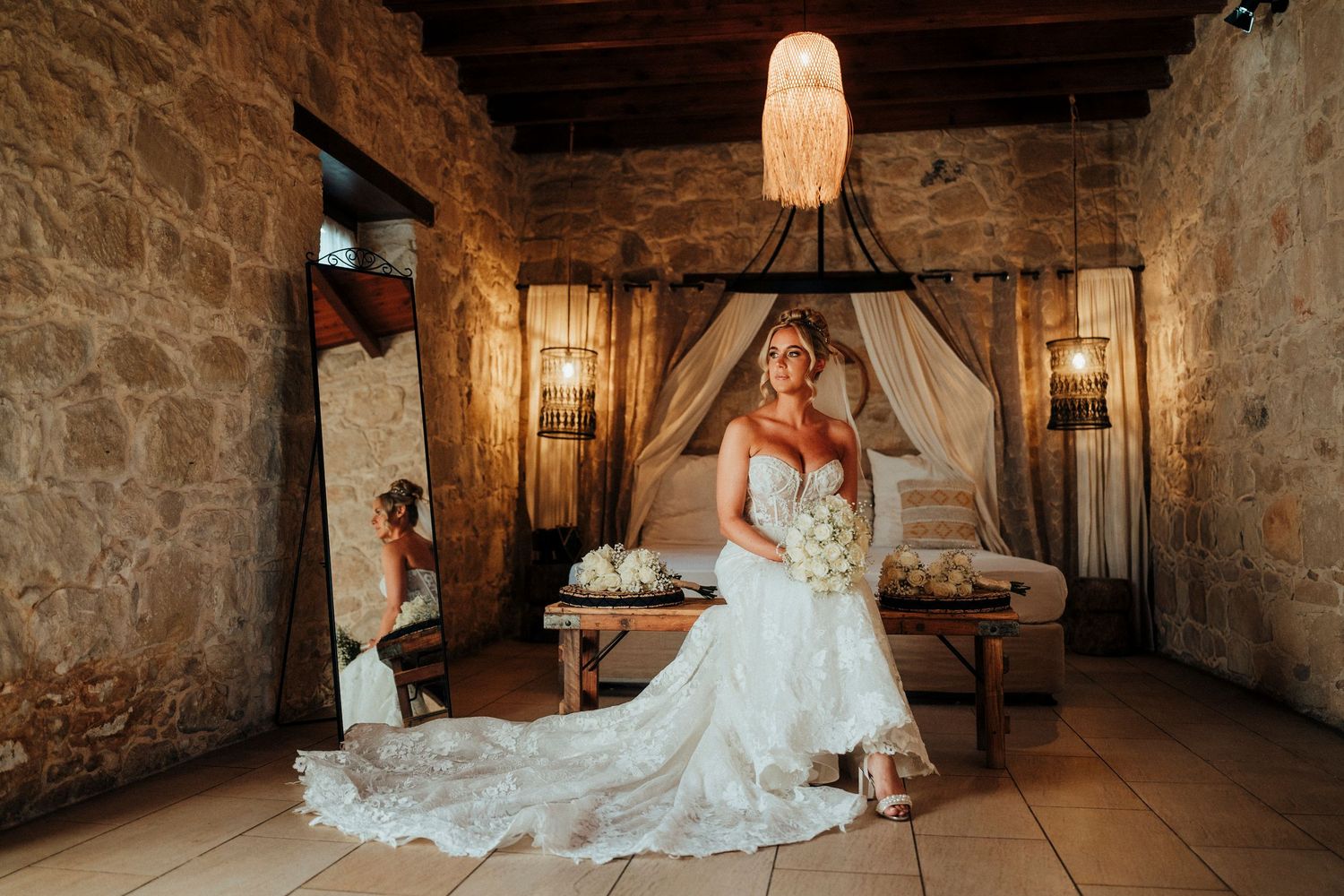A bride in a white lace dress stands by a window holding a bouquet.