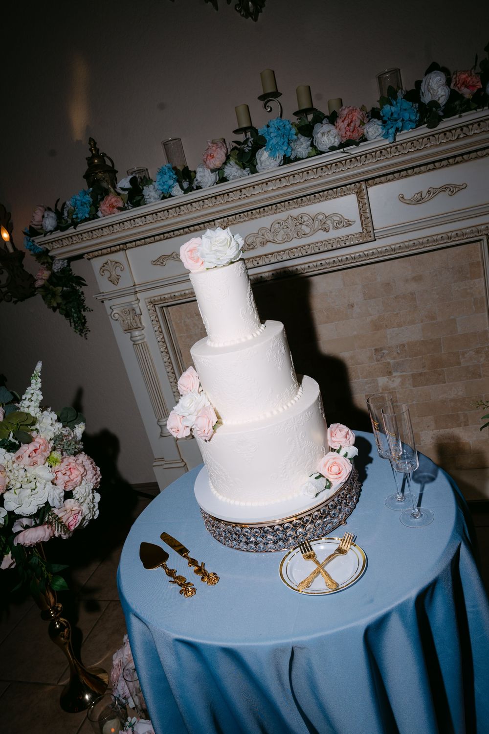 Elegant four-tier white wedding cake with pink roses on a silver stand, set against a floral fireplace backdrop.