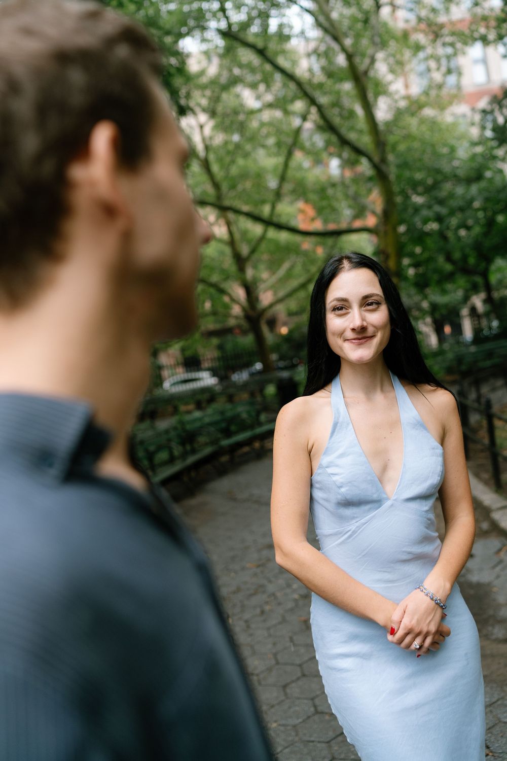 A smiling woman in a light blue halter dress stands outdoors in a park, facing a man in a dark shirt.