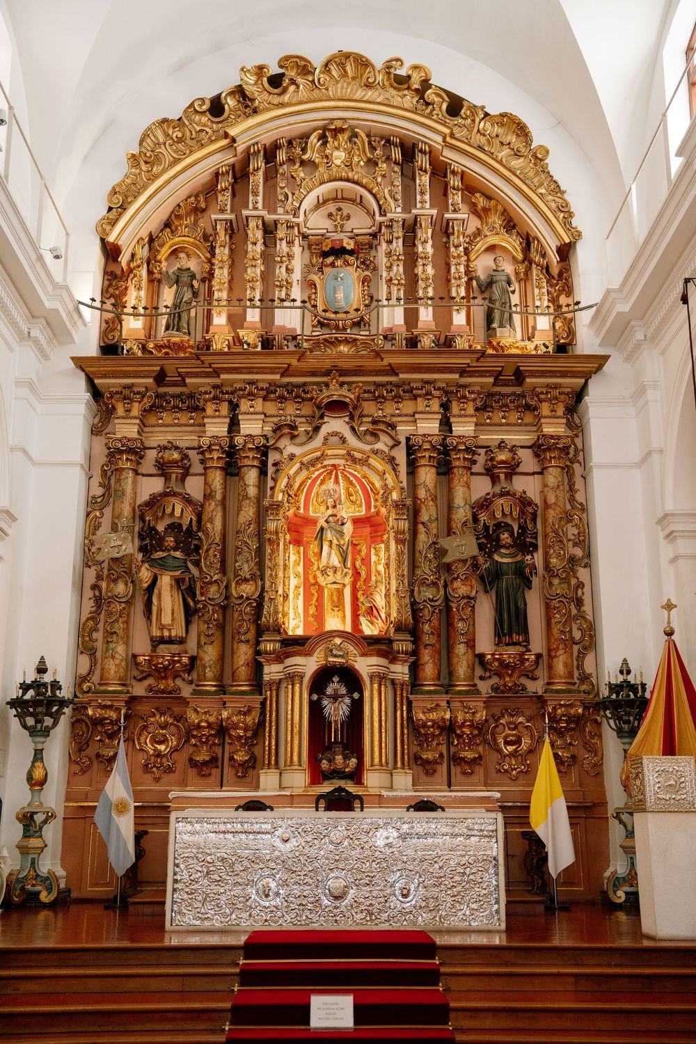 Ornate golden baroque altar with religious statues in a white church, with Argentine and Vatican flags beside it.