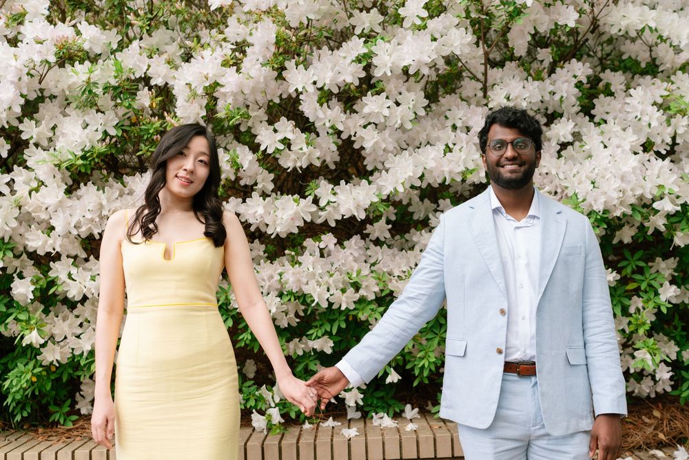 A couple poses together in front of a blooming white floral backdrop in a garden setting.