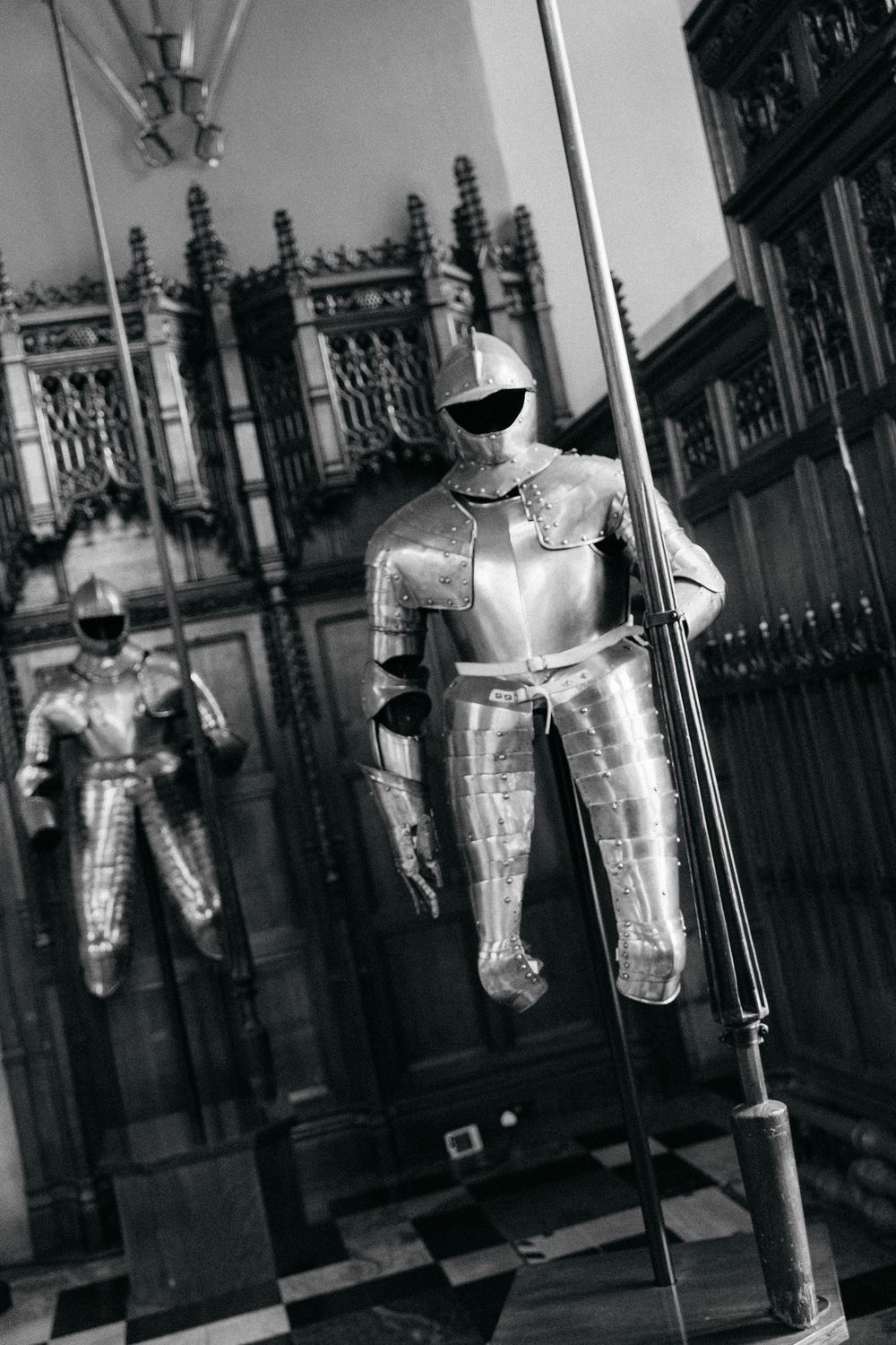 Black and white photo of two polished medieval knight armor suits on display in a historic castle interior.