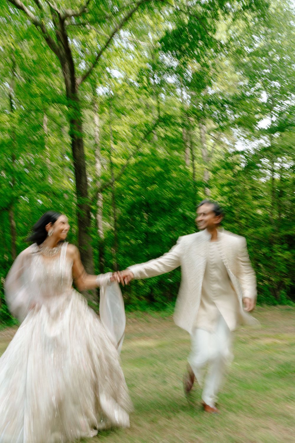Blurred motion photo of Indian bride and groom holding hands running through green forest.