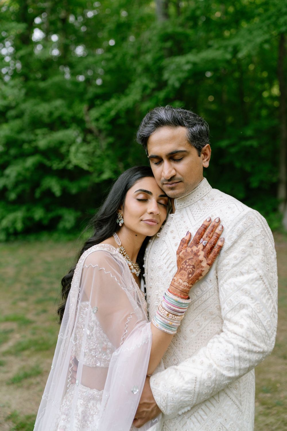 Indian bride with mehndi hands embracing groom in white sherwani outdoors in forest.