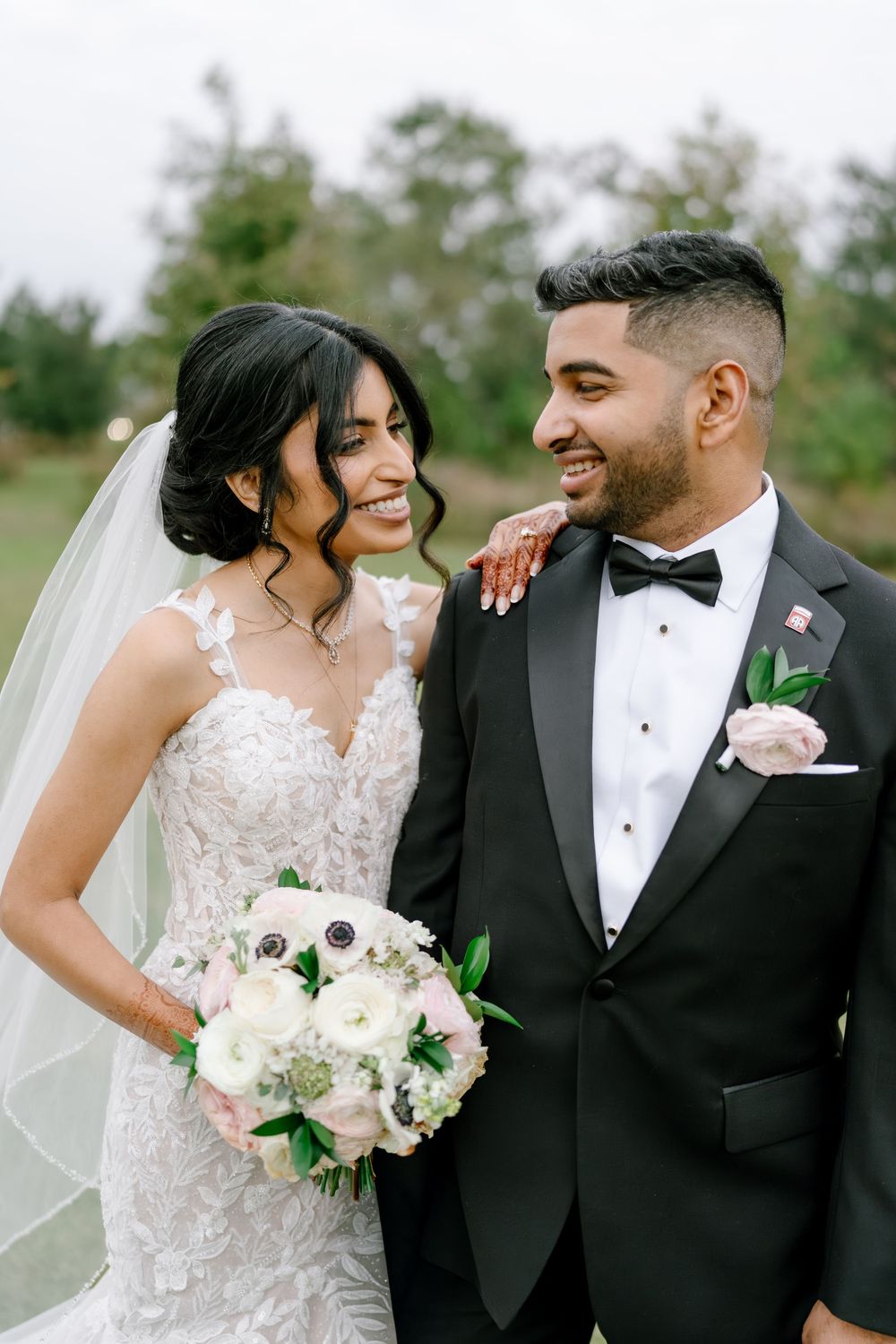 A bride and groom smile at each other outdoors, she holds a white bouquet and wears lace gown, he wears black tuxedo. Houston Texas Wedding.