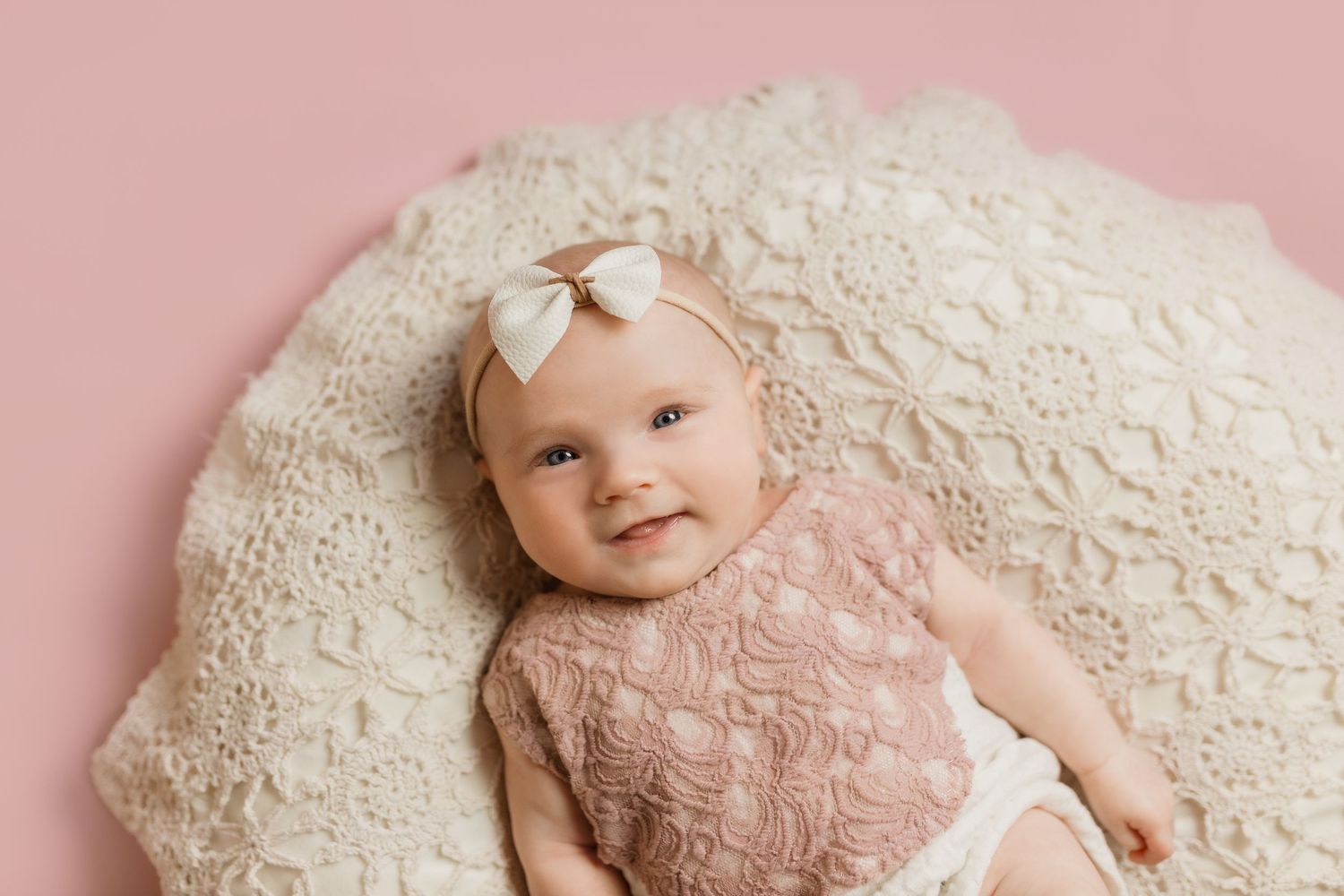 Baby wearing pink lace dress and bow lying on cream textured blanket with soft pink background.