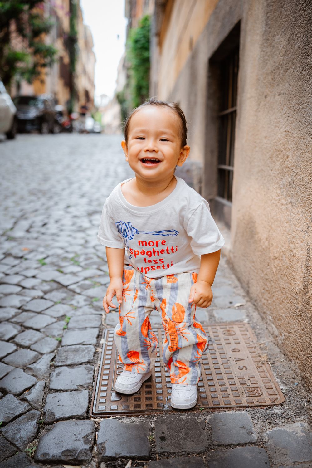 Smiling toddler in spaghetti print outfit standing on cobblestone street in Rome, Italy.
