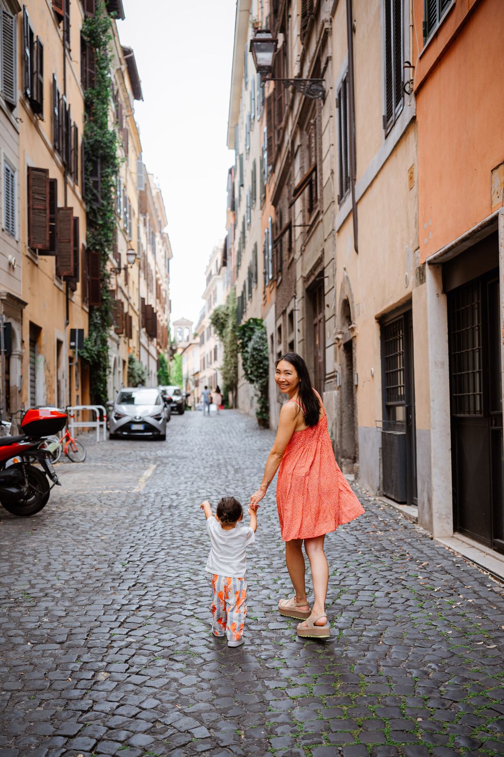 Mother in coral dress holds toddlers hand walking on cobblestone street in Rome.