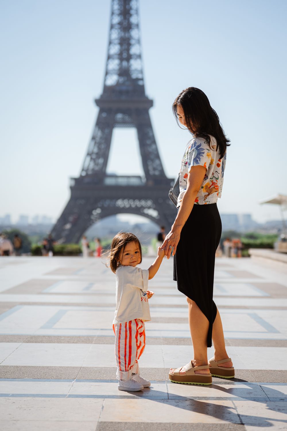 A mother and young daughter hold hands at Trocadero plaza with the Eiffel Tower in Paris, France behind them.