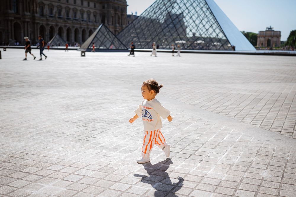 A young child runs joyfully across the Louvre plaza in Paris with the iconic glass pyramid in the background.