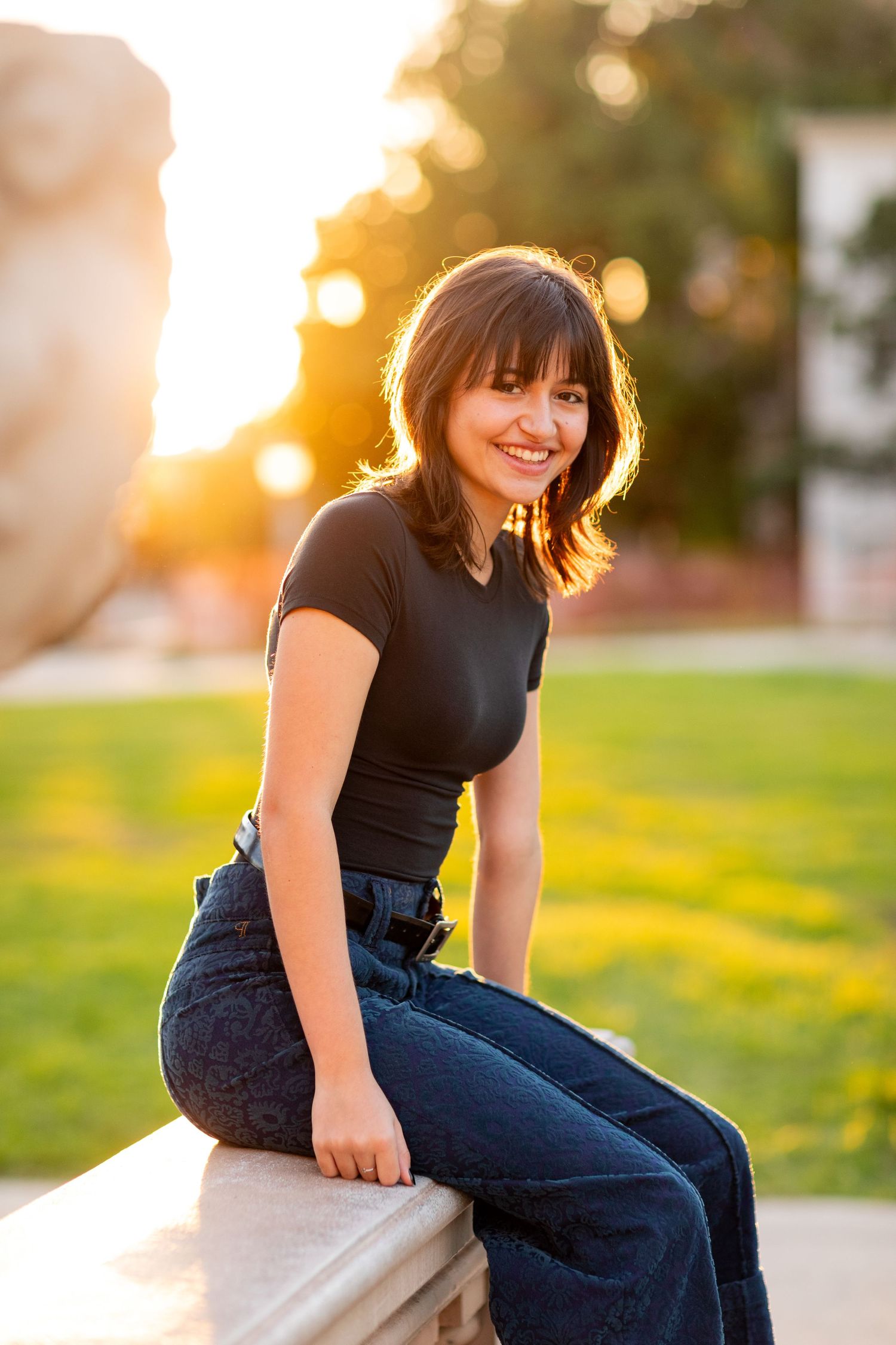 A high school senior in black shirt and jeans sits outdoors at sunset with golden bokeh lighting in the background.