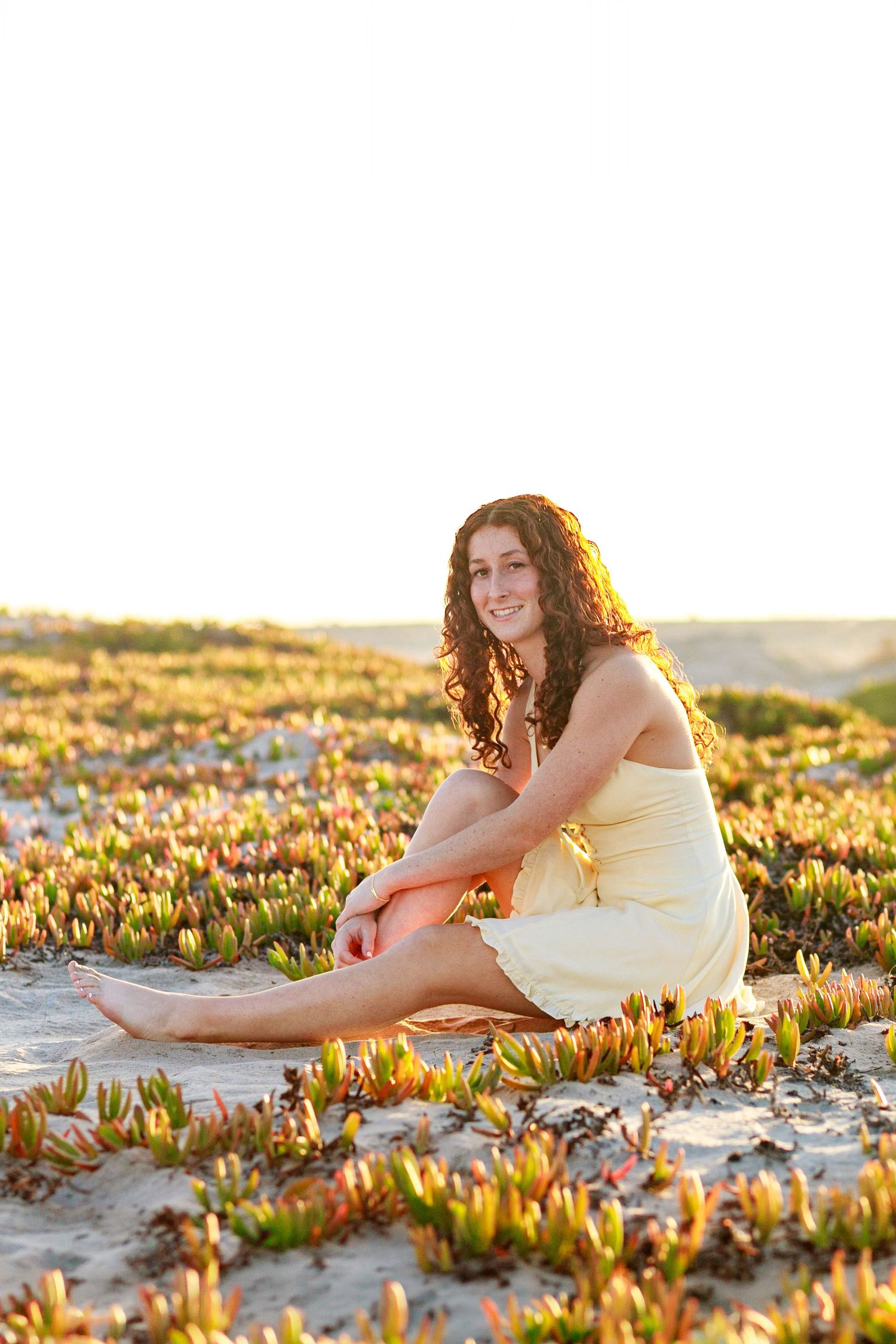 A high school senior in black shirt and jeans sits outdoors at sunset with golden bokeh lighting in the background.
