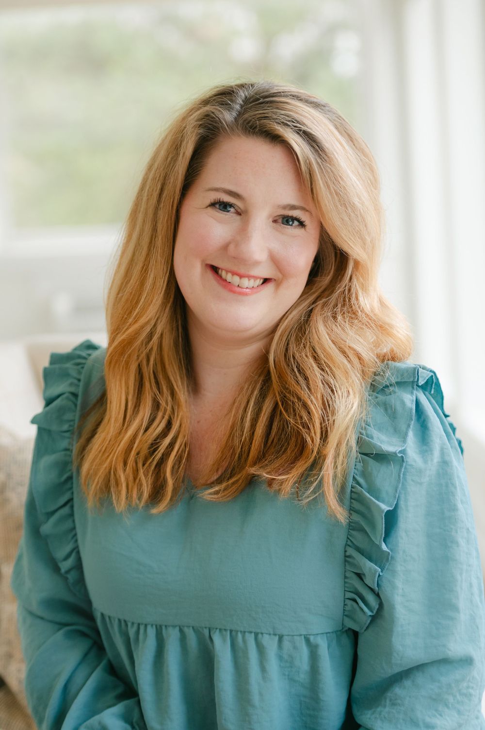 Smiling woman with blonde hair wearing a teal ruffled blouse indoors.