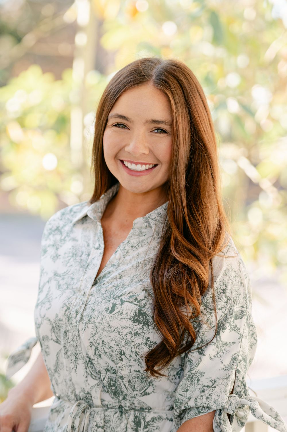 Young woman with auburn hair smiling outdoors in a floral green and white dress.