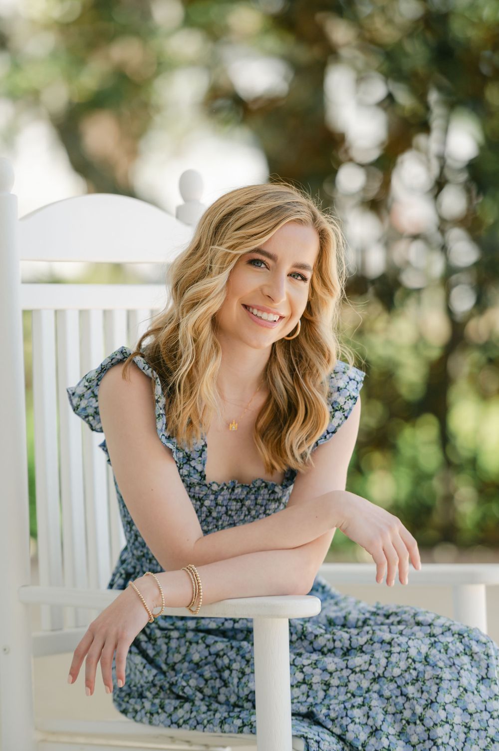 Blonde woman in a blue floral dress sitting on a white bench outdoors smiling.