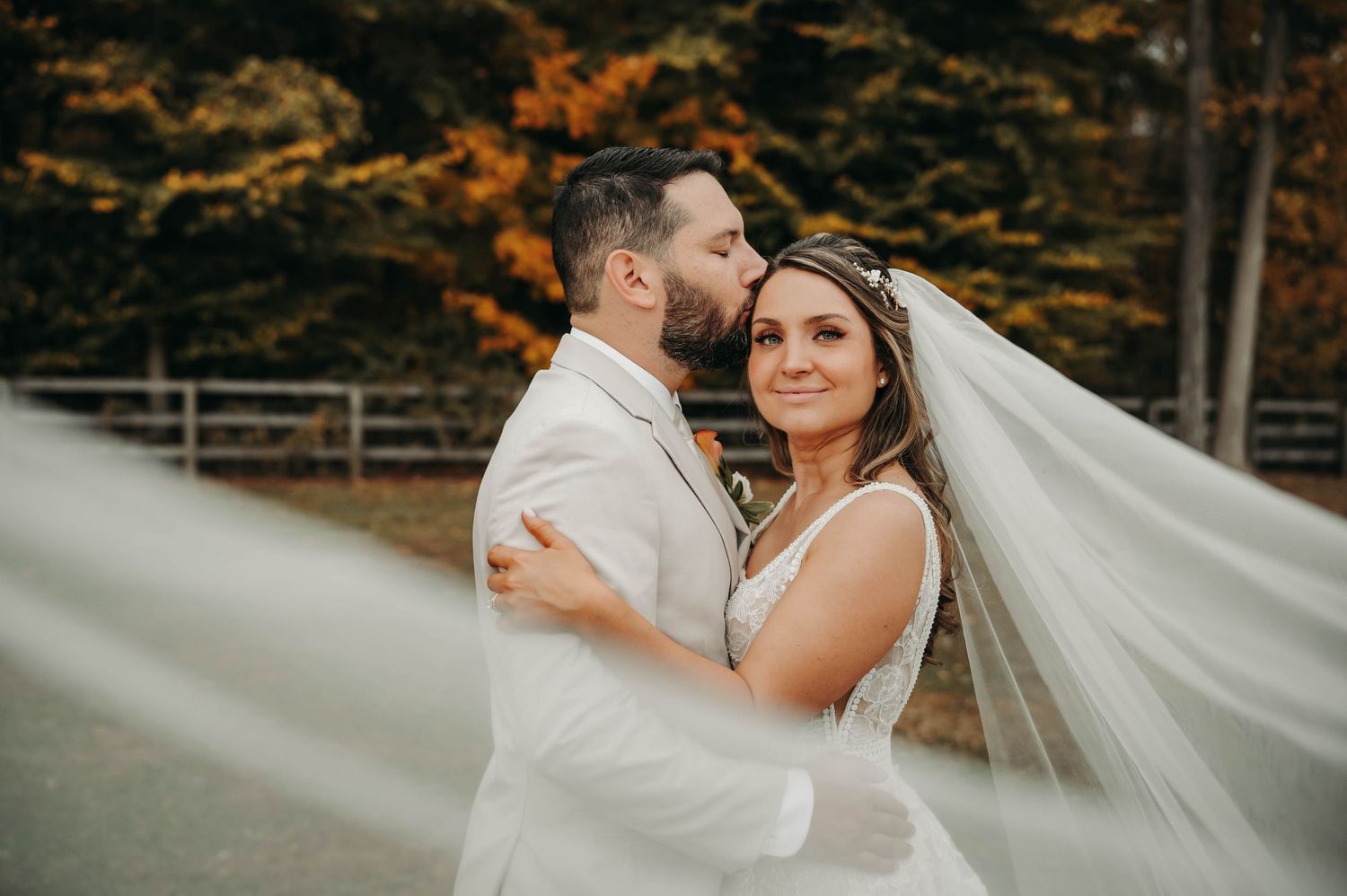 A bride smiles as her groom kisses her cheek while her veil flows in the autumn breeze.