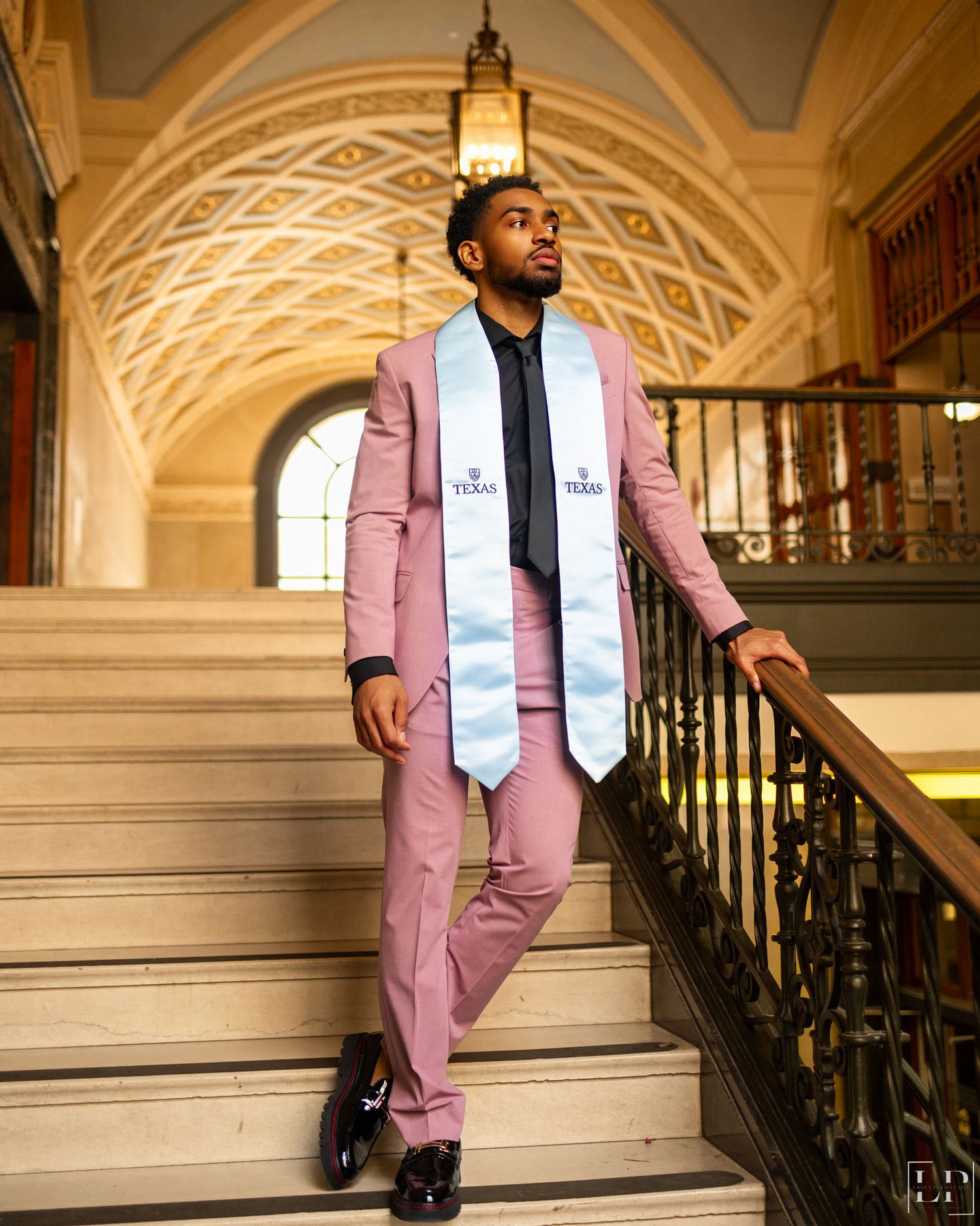 Graduate in pink suit poses on ornate staircase with classical architecture.