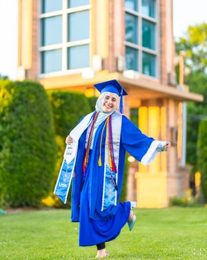 Graduate in blue cap and gown celebrating on sunny campus lawn.