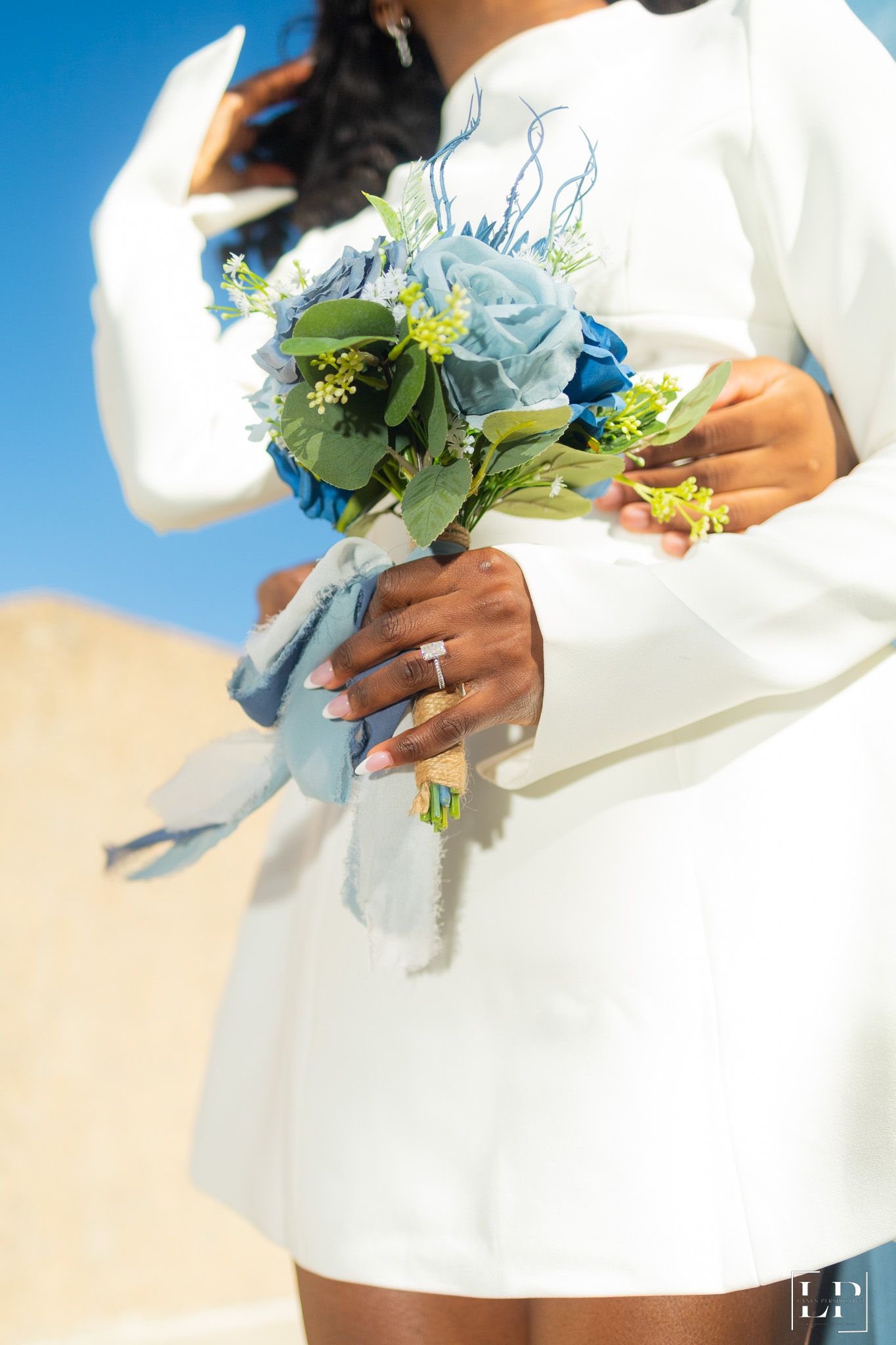 Close-up of white wedding dress and blue floral bouquet details against sunny backdrop.