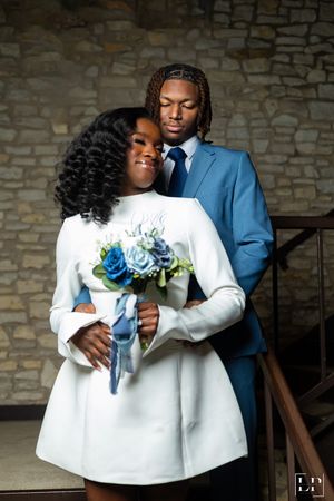 Couple in formal attire holding blue and white bouquet against stone wall background.