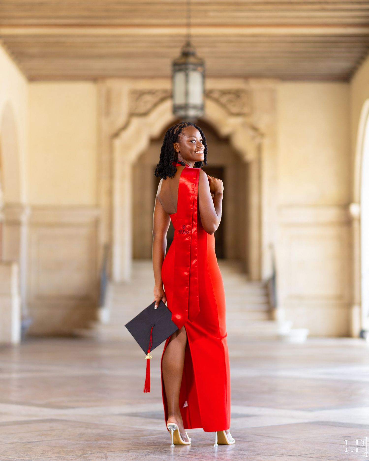 Graduate in vibrant red dress poses elegantly in columned hallway.