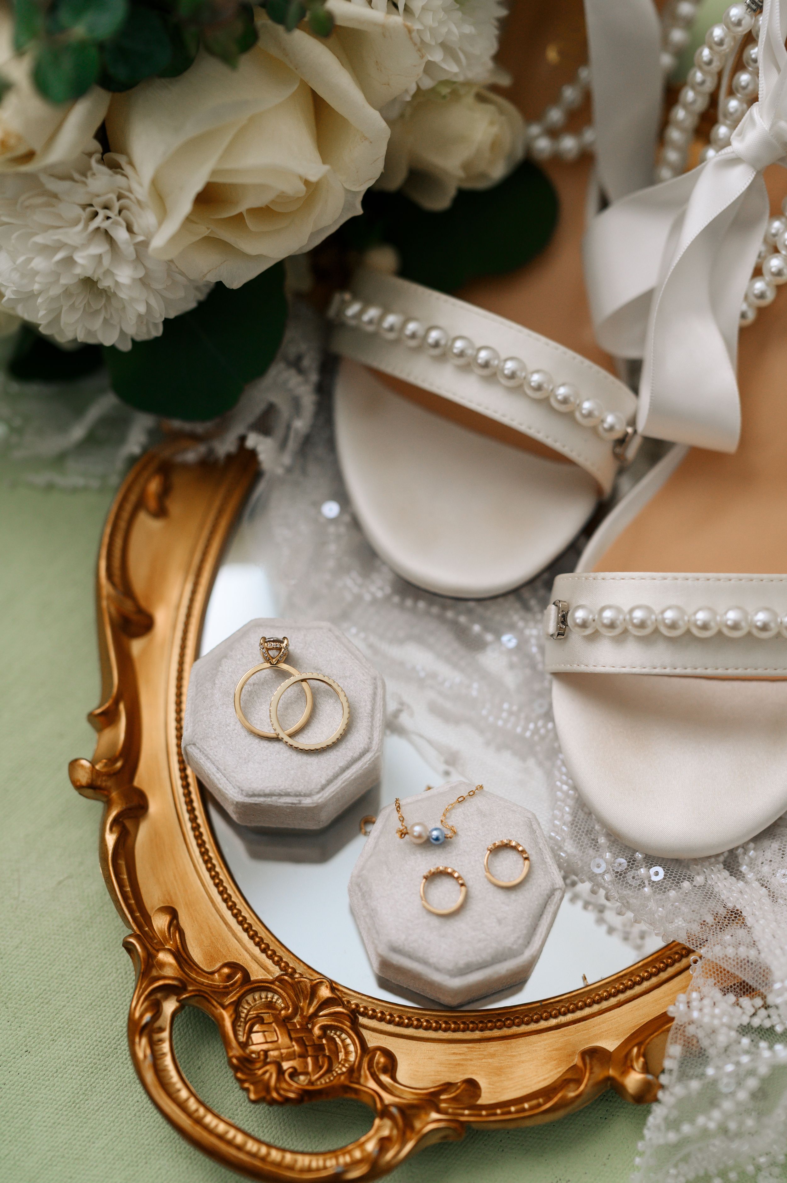 Wedding accessories including pearl necklace, gold rings, and white heels arranged on an ornate gold mirror with floral details.