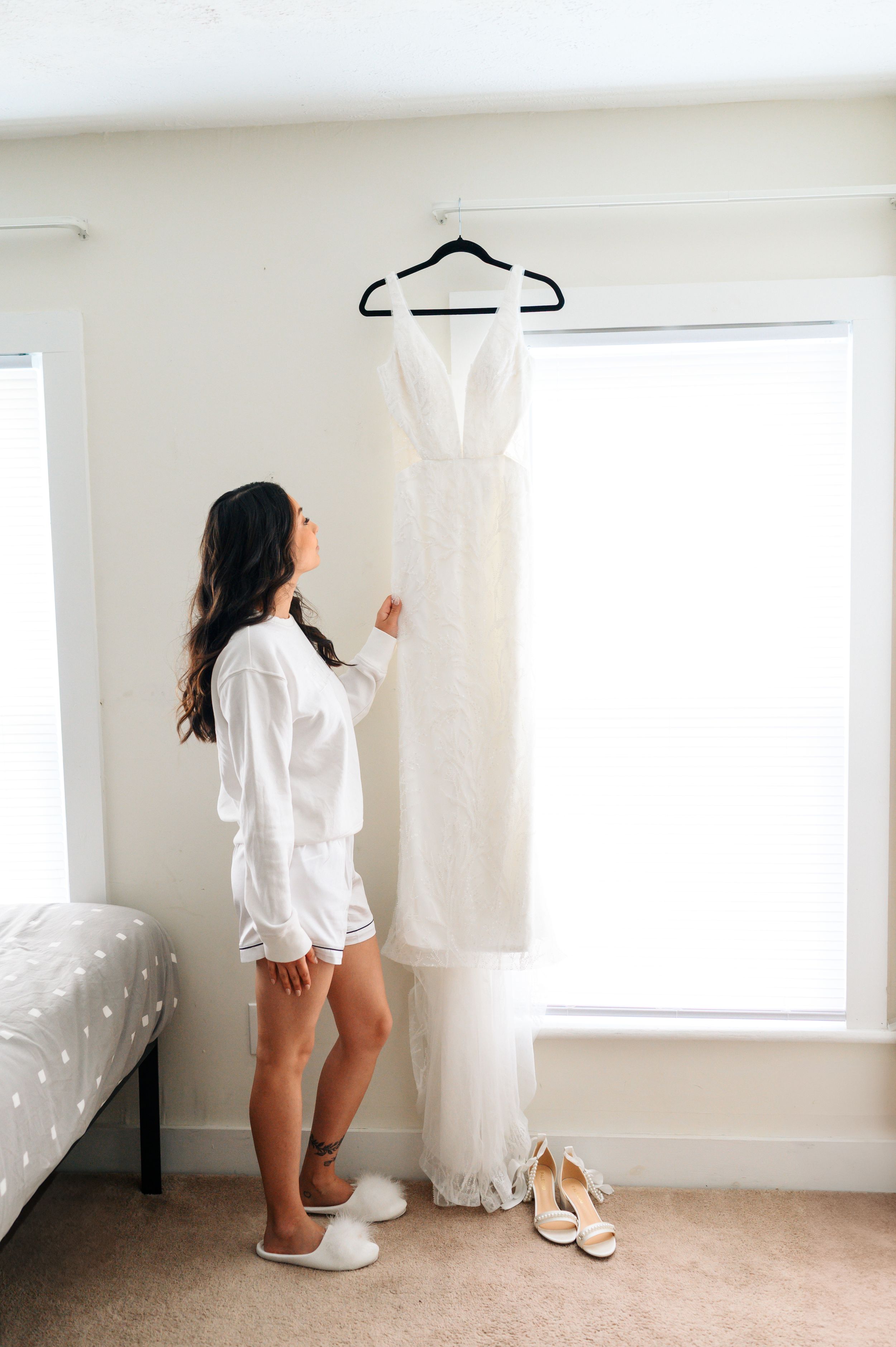 A white wedding dress hangs in a bright room while someone in a white robe admires it next to beige heels.