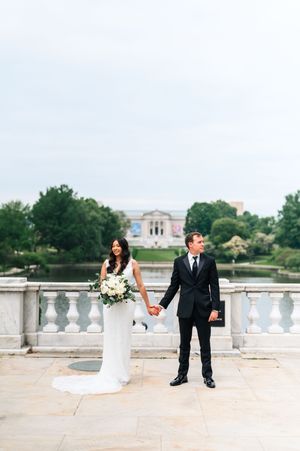 A wedding couple poses on a terrace overlooking a classical building and lake in the background.
