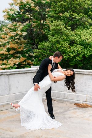 A couple in wedding attire performs a romantic dip pose on a stone terrace surrounded by lush green trees.