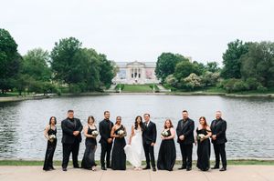 Wedding party in black formal attire poses by a serene lake with a grand building in the background.