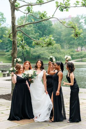 Bridesmaids in black dresses and bride in white gown pose playfully by a lake for wedding photos.