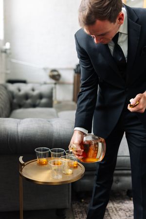 Person in dark suit pouring whiskey into glasses on a round wooden side table in a modern living room setting.