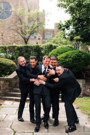 Group in formal black suits posing playfully outdoors against a stone building with greenery.