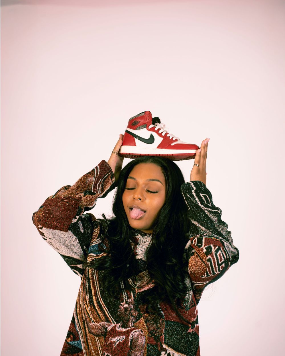 A person in a brown leather jacket poses playfully with a red Nike sneaker against a pink background.