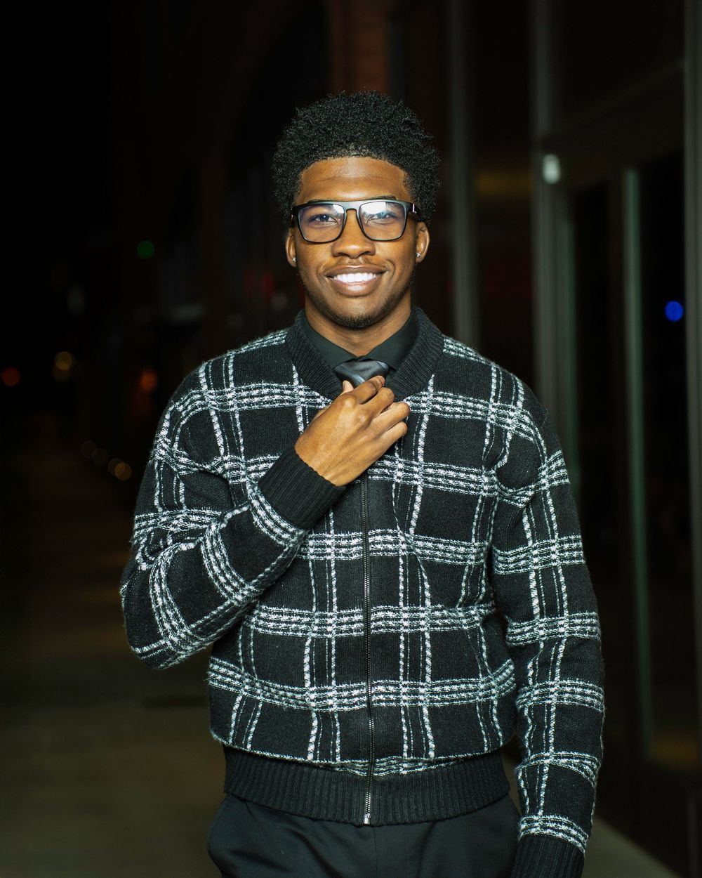 Someone wearing glasses and a black and white plaid sweater smiles while gesturing at night.