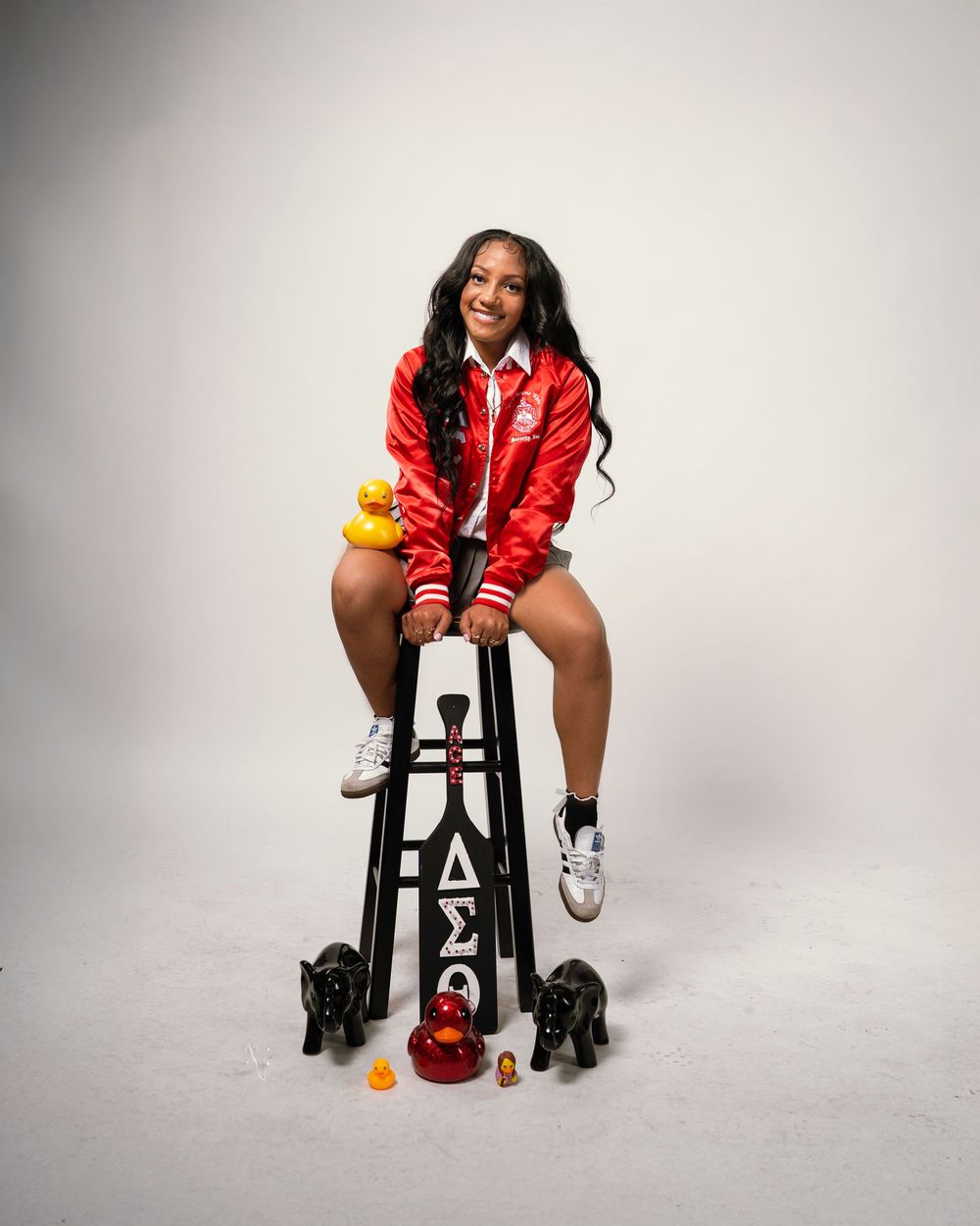 A model in a red varsity jacket and white sneakers poses on a stool against a white studio backdrop.