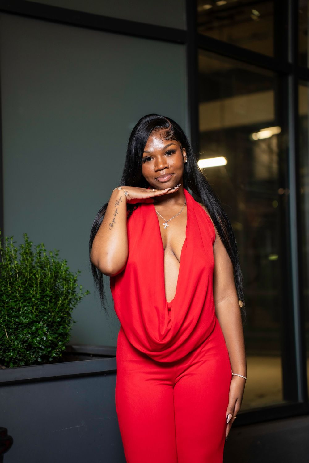 A woman in a vibrant red jumpsuit poses confidently against a dark background with greenery visible.