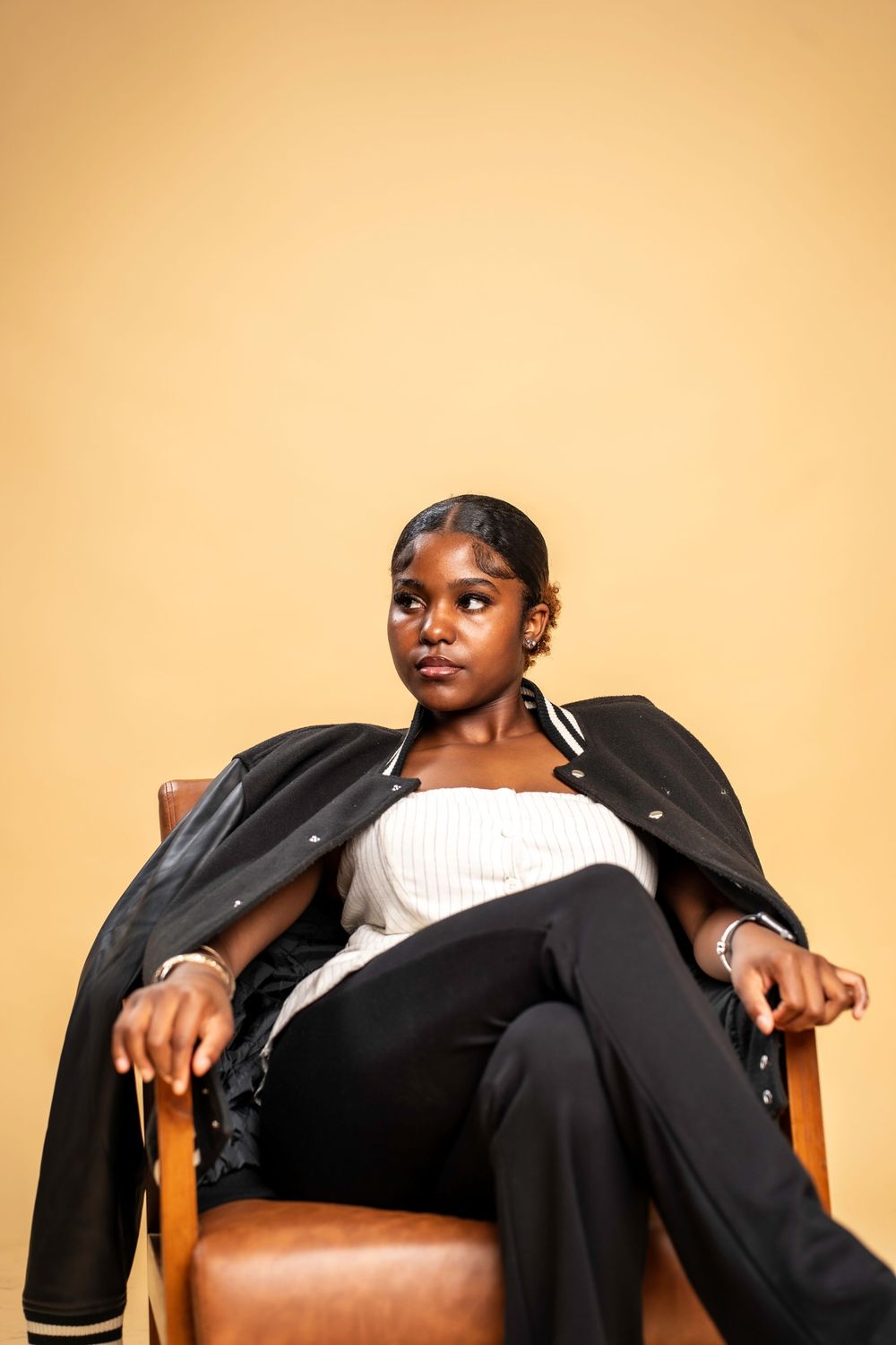 A person in a black blazer and white top reclines on a wooden chair against an orange background.