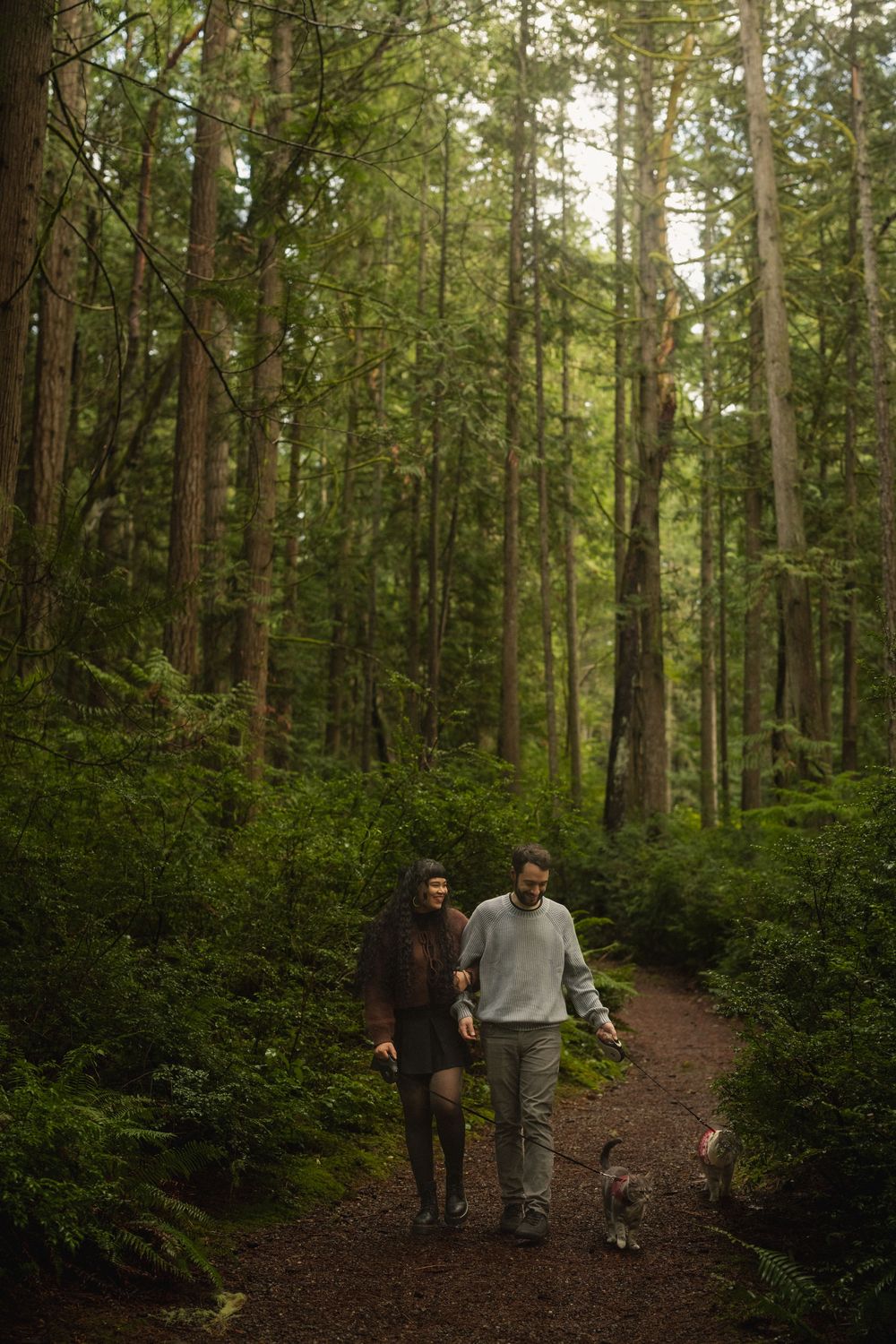 Two people walk together on a hiking trail through a lush green forest of tall evergreen trees.