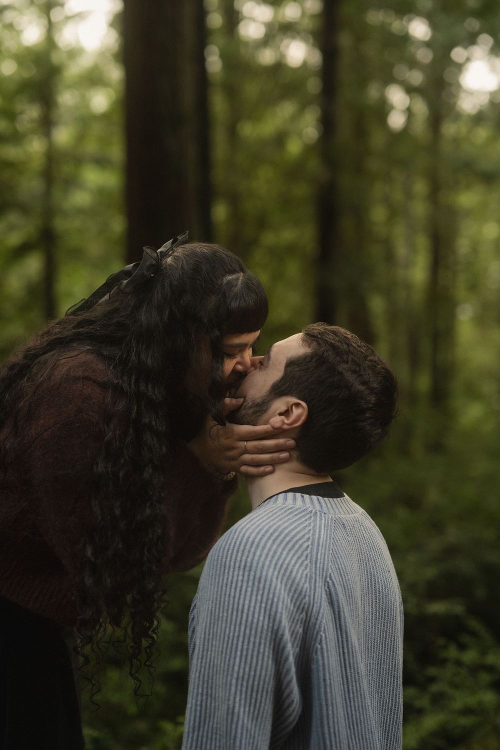 A couple shares a romantic kiss in a lush green forest setting.