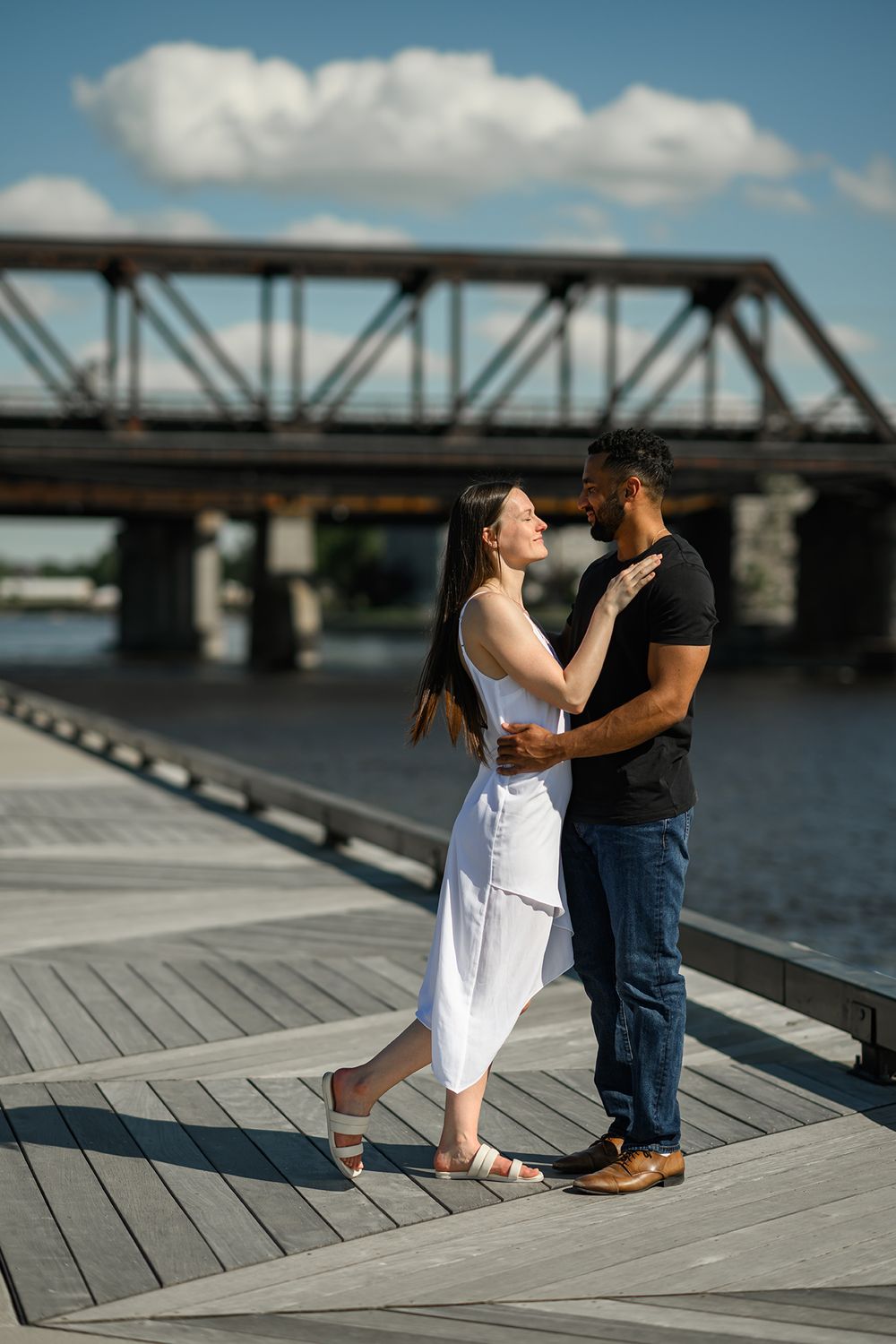 A couple in casual attire sharing an intimate moment on a wooden deck with a steel bridge in the background.