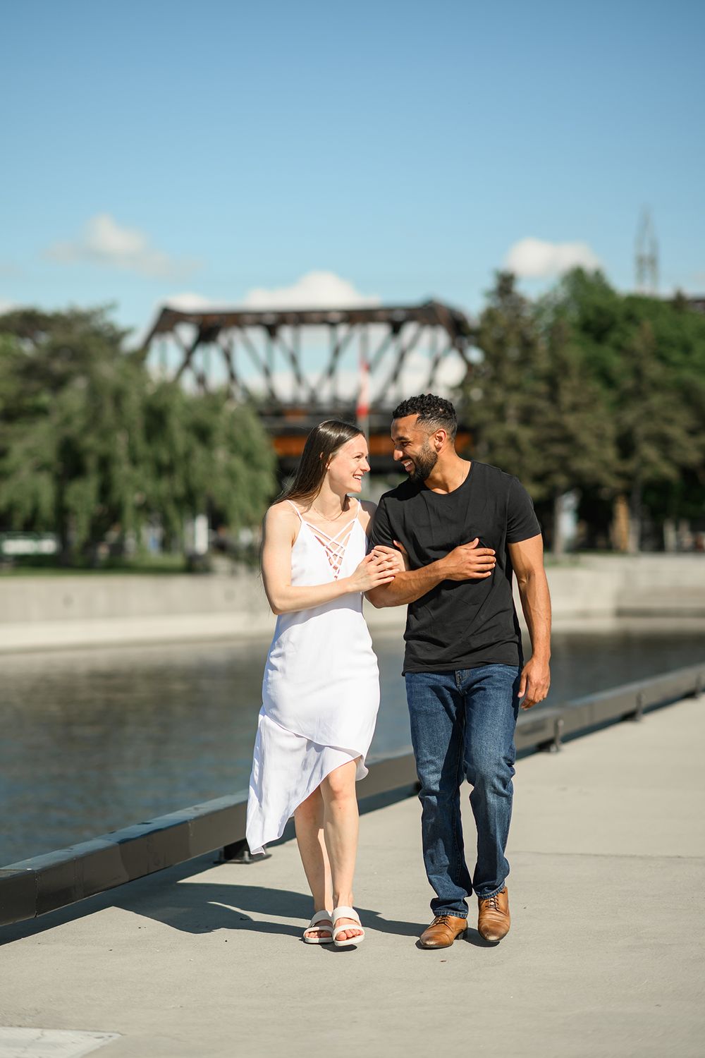 A couple shares a romantic moment sitting on a concrete walkway near a bridge on a sunny afternoon.