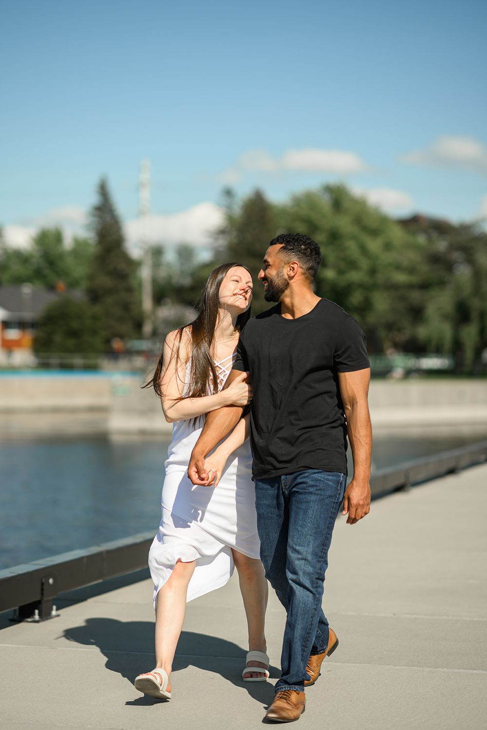 A couple shares laughs while walking along a waterfront promenade on a sunny afternoon.