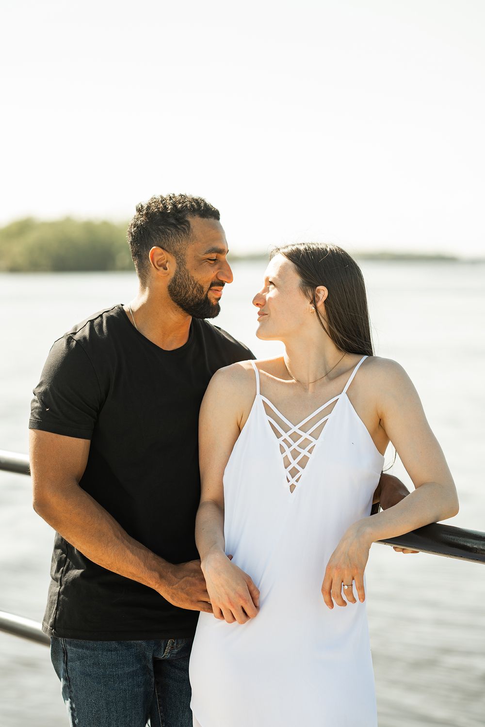A romantic couple in black and white outfits share intimate moments by the water during a photoshoot.